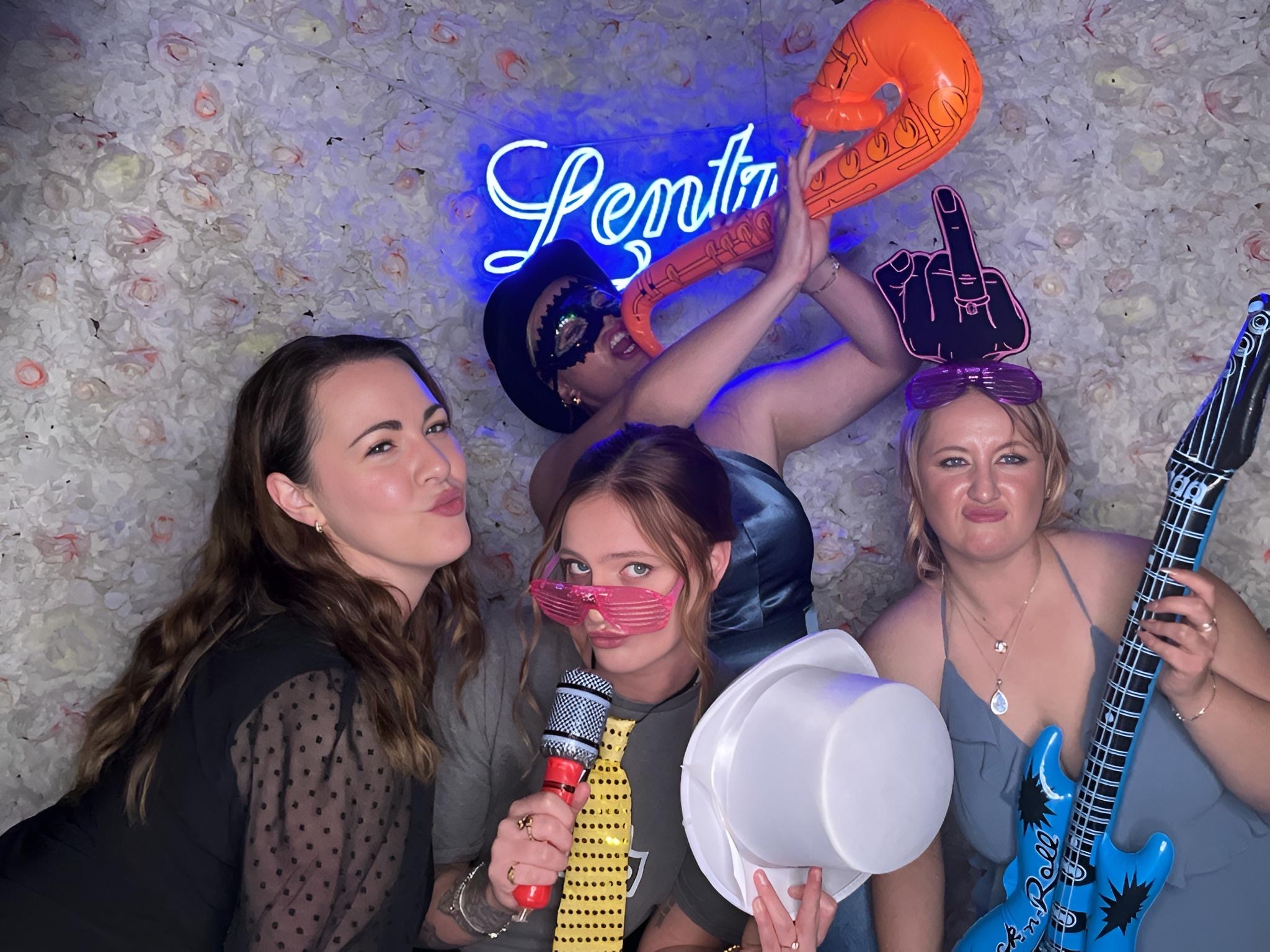 "Group of women posing with inflatable props and hats in front of a flower wall and neon sign at a photo booth event"