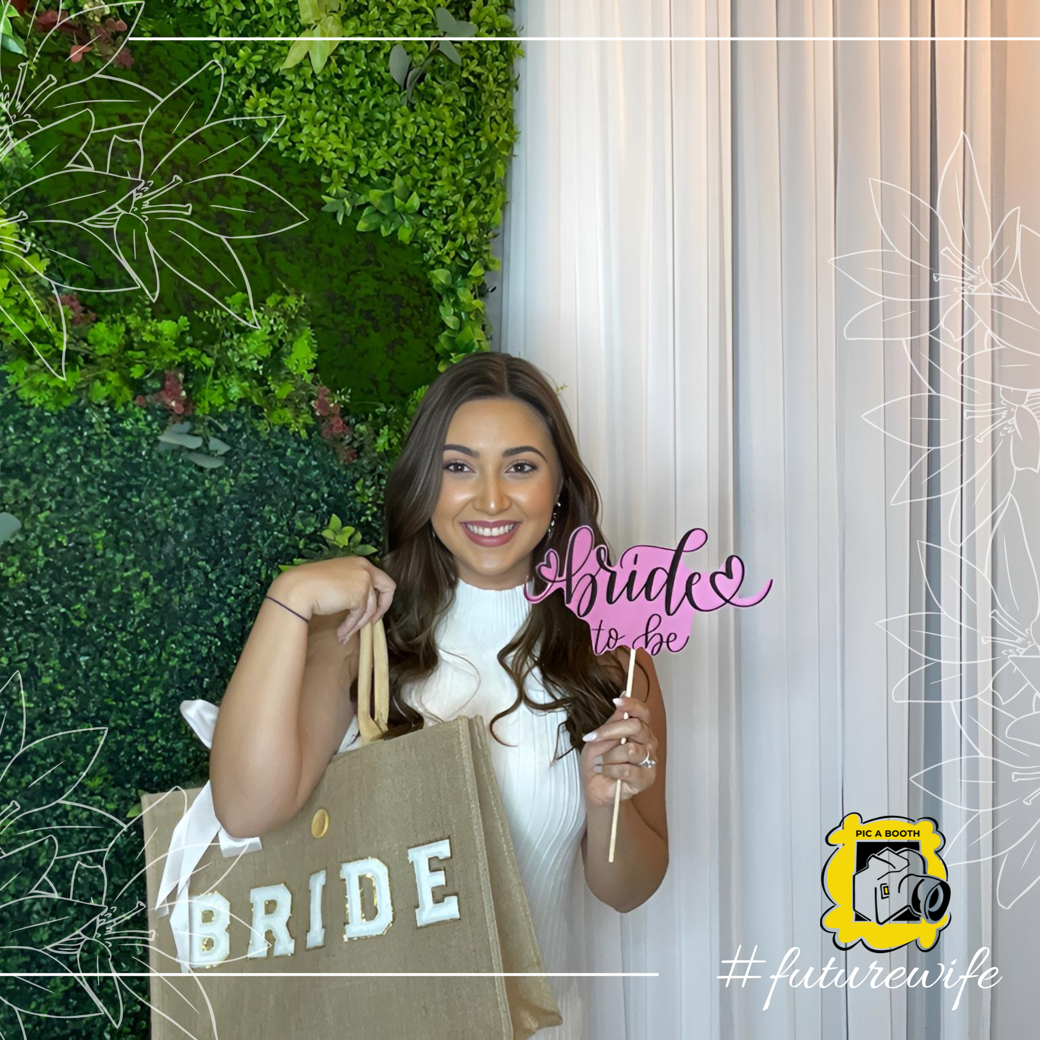 "Smiling bride-to-be holding bridal props and tote bag at PIC-A-BOOTH photo booth with greenery and drape backdrop"